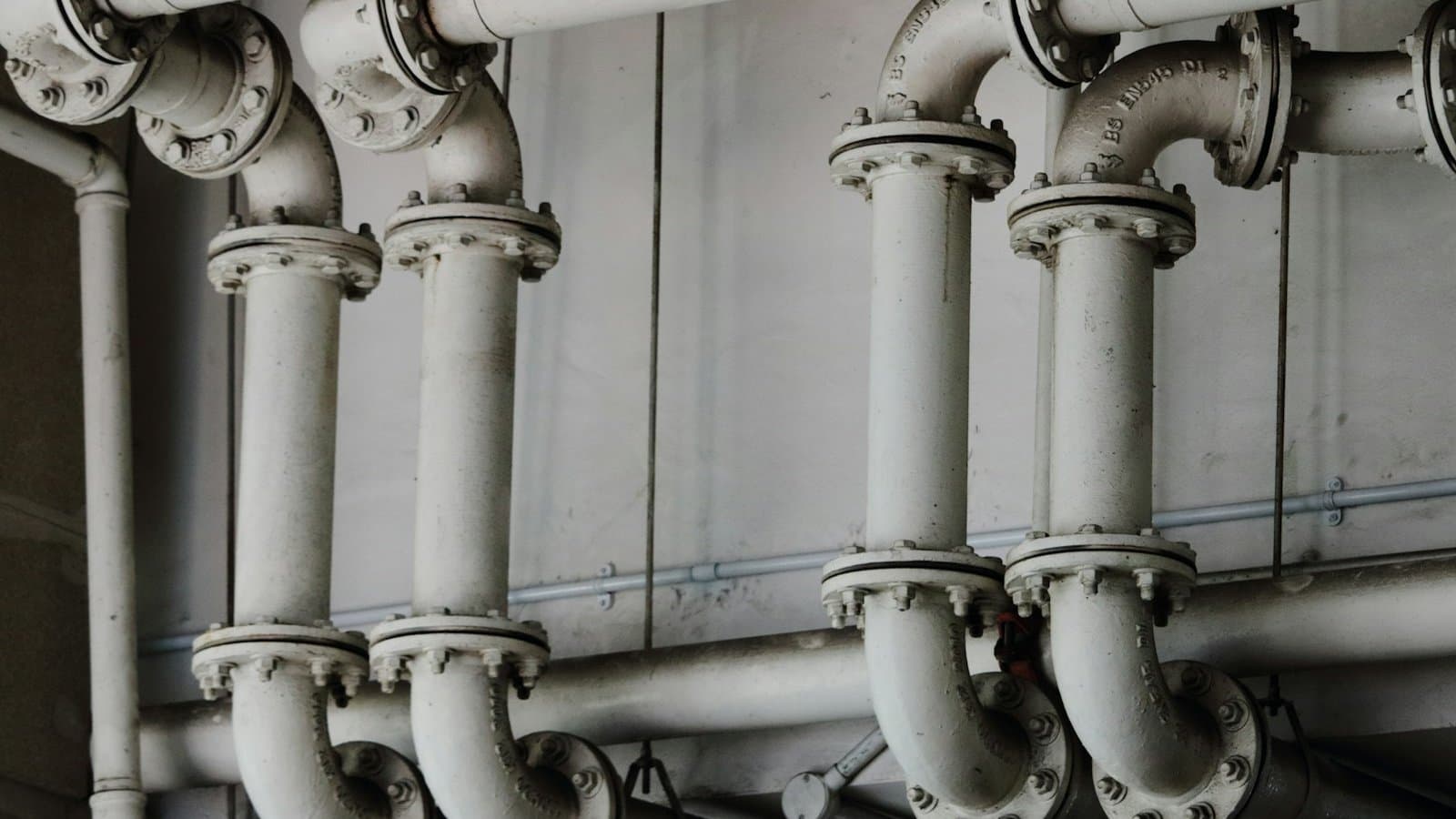 Grayscale close-up of industrial metal pipes, the building water system pipework that carries Legionella risk when water temperatures and stagnation create aerosol exposure