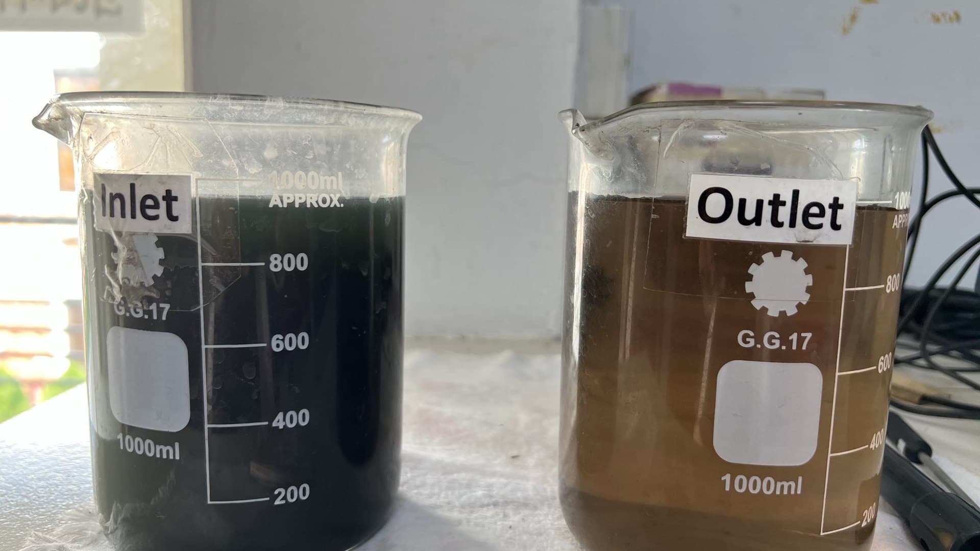Labelled inlet and outlet beakers from a textile dyehouse effluent plant — near-black dye liquor beside the decolourised treated water
